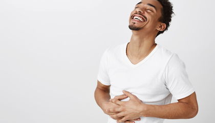 Smiling man in a white t-shirt laughing with eyes closed, hands resting on his stomach.