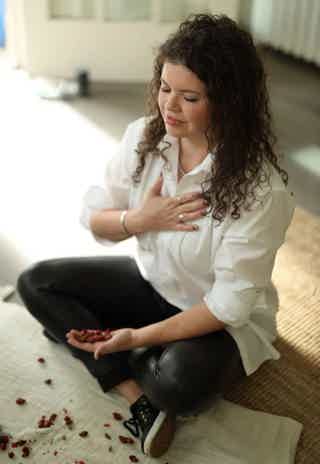 Woman sitting cross-legged on rug, eyes closed, hand on chest, petals scattered nearby.