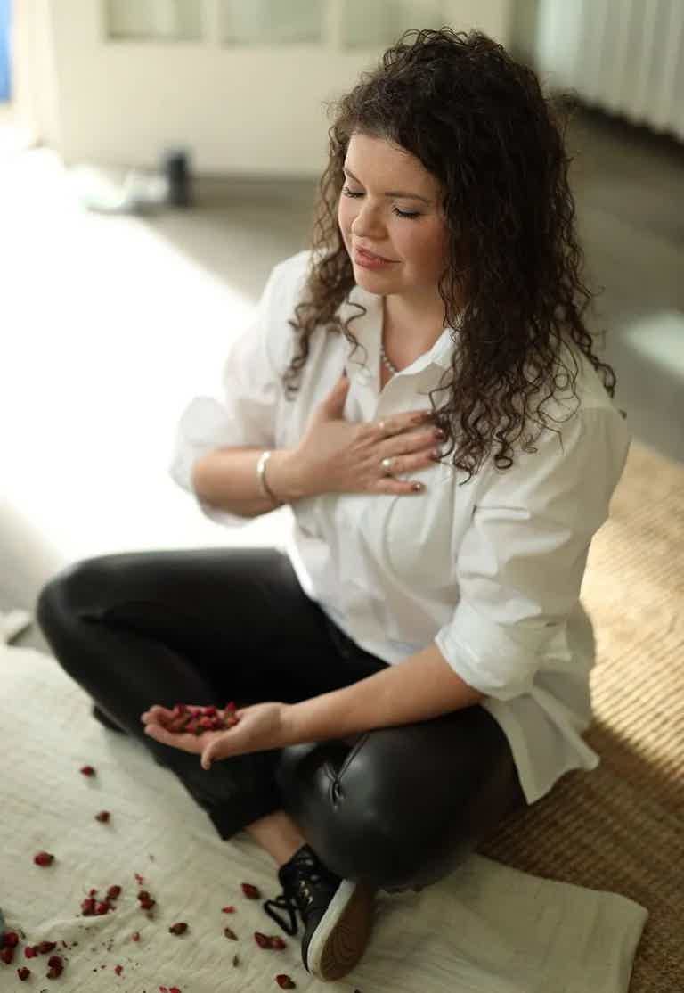 Woman sitting cross-legged on rug, eyes closed, hand on chest, petals scattered nearby.