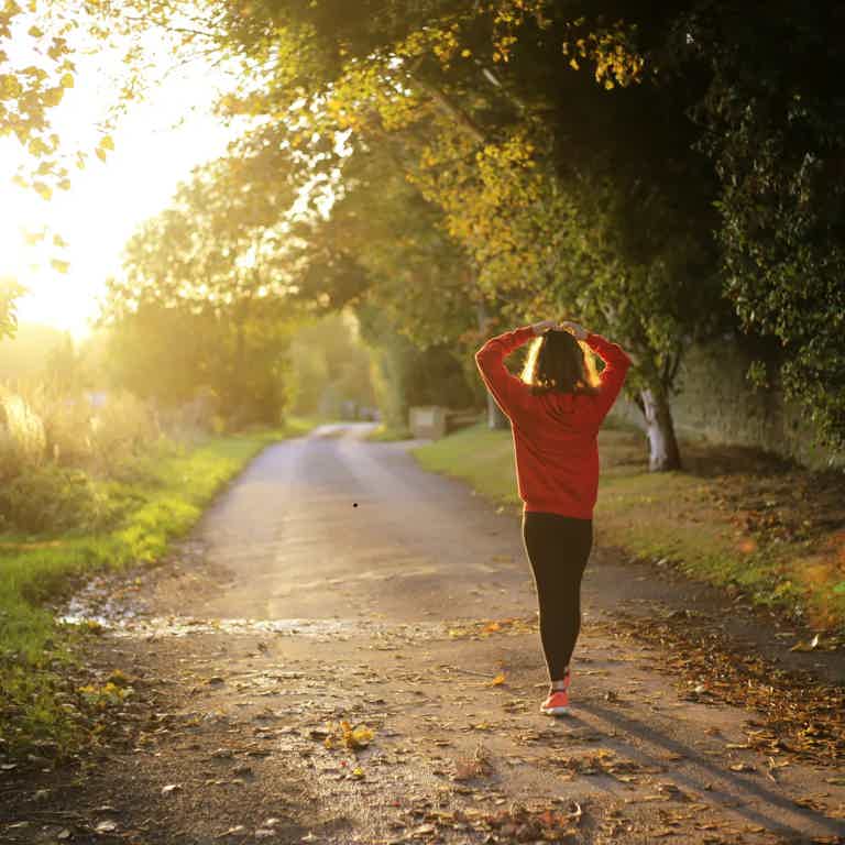 Person in red hoodie stands on a sunlit country road, hands behind head, trees lining path with autumn leaves.