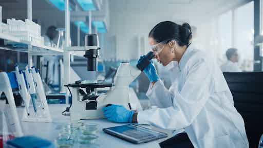 Female scientist in a white lab coat and blue gloves examining a microscope slide in a modern clinical laboratory.