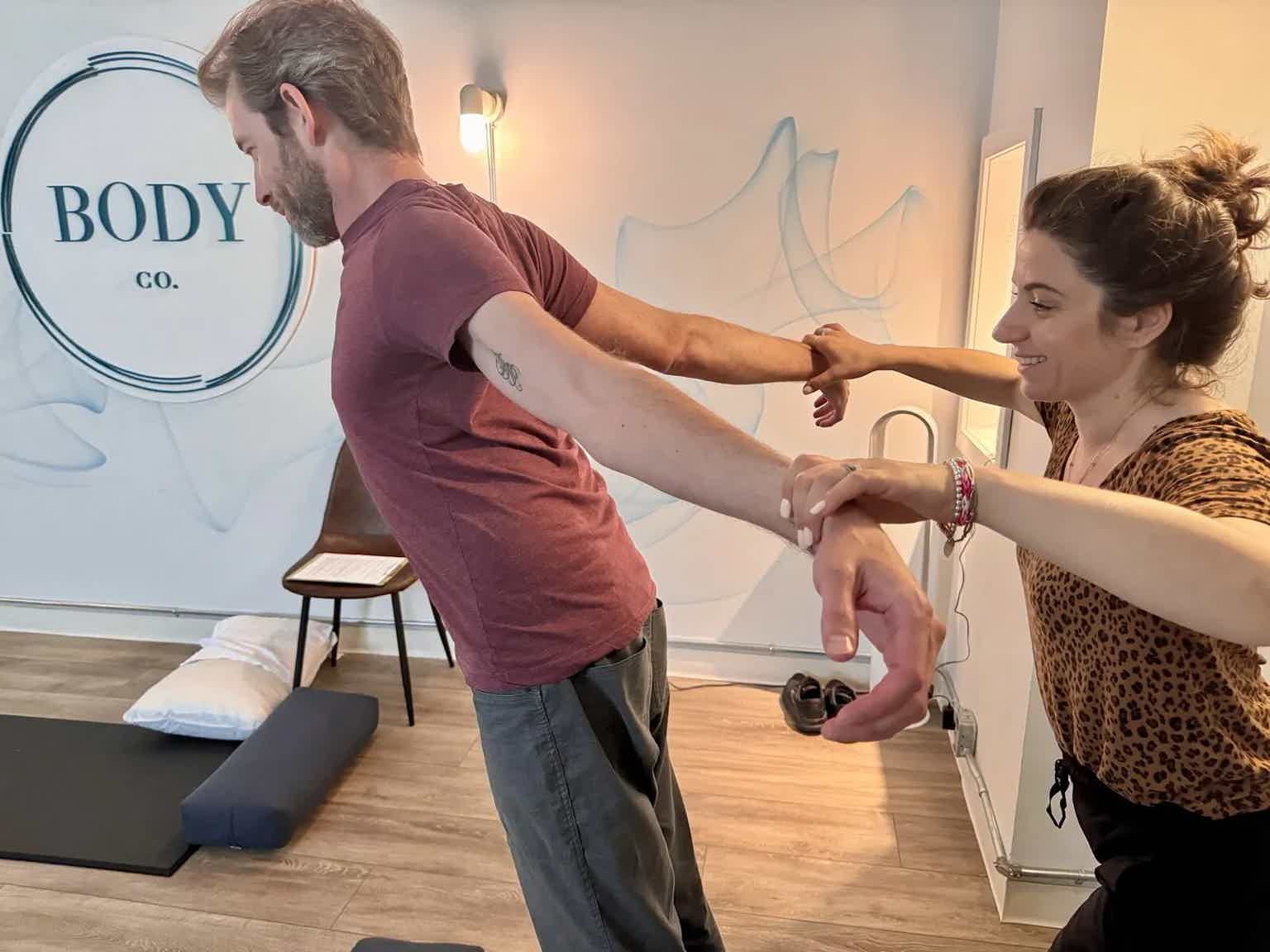 Instructor and participant stretch arms at bodywork studio, with wall signage “BODY CO.” and a bench in background.