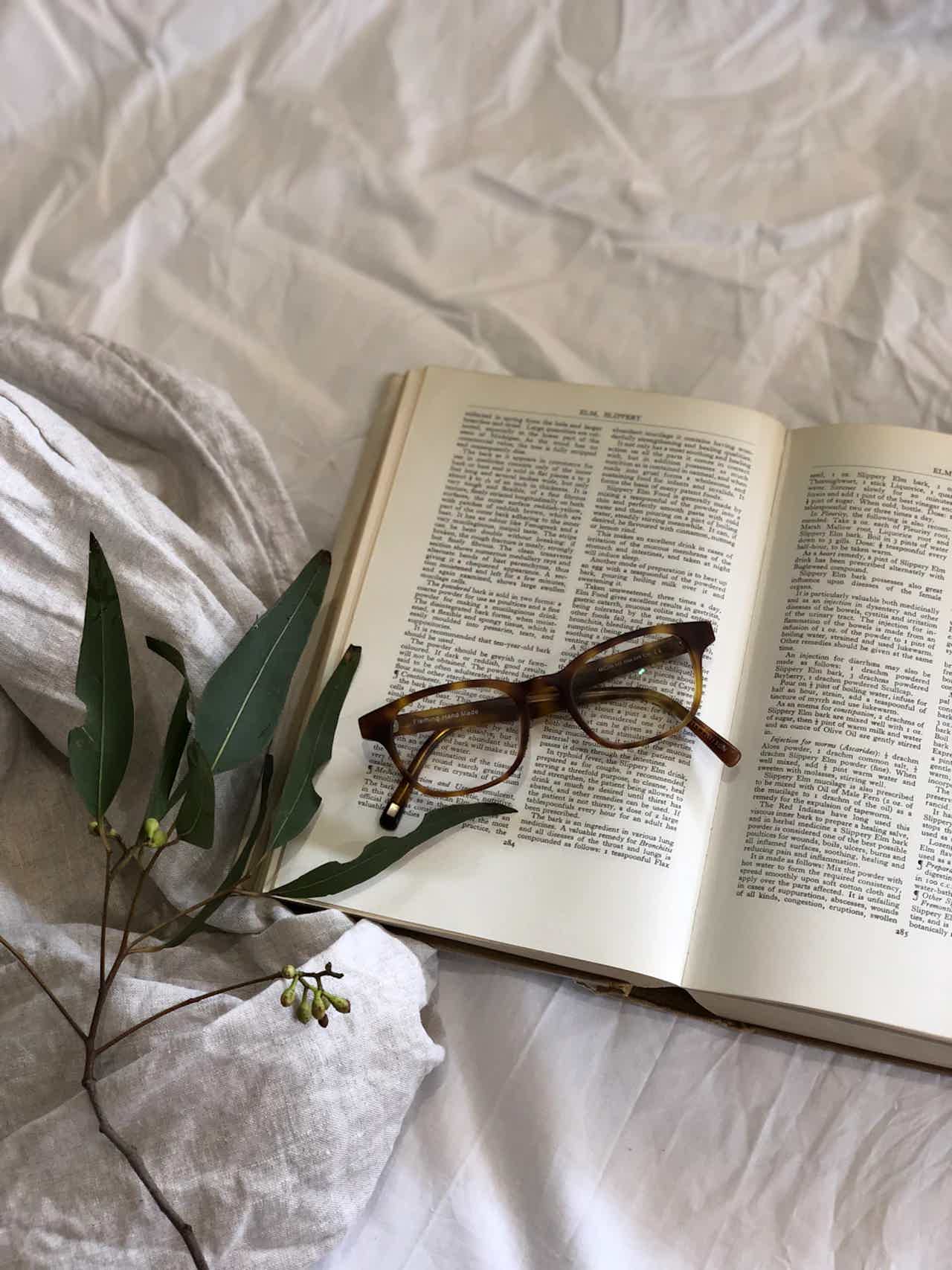 Open book with text, brown eyeglasses, and green leaves on crumpled white fabric background.