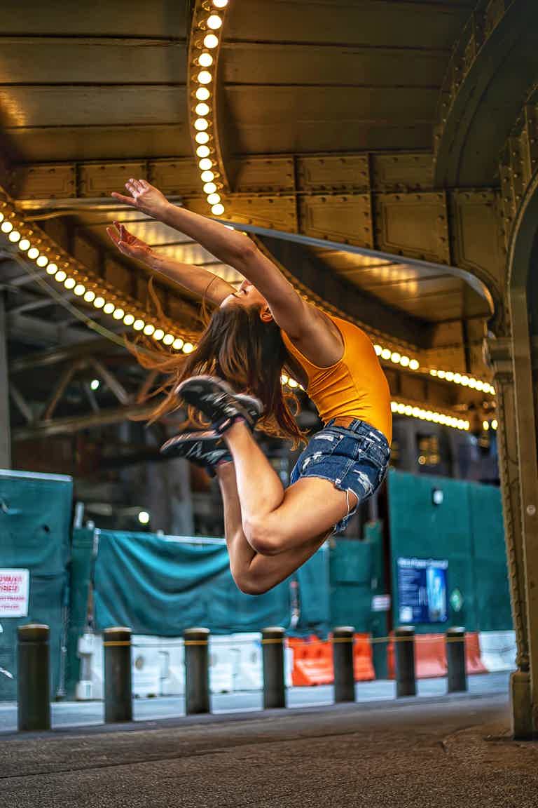 Two people mid-air jump under a lit city bridge, arms extended, cheering on a nighttime urban boardwalk.