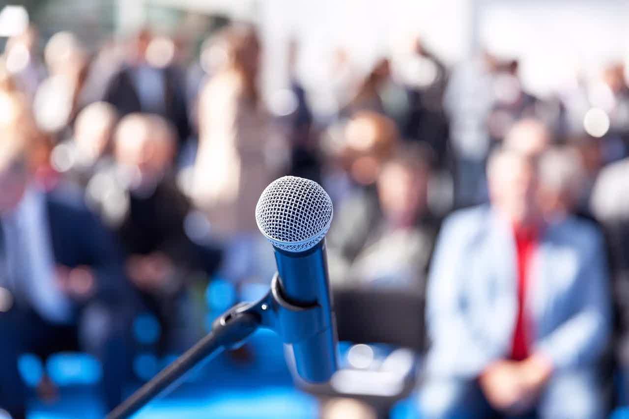 Microphone on stand foreground with blurred audience in a conference or event setting behind.