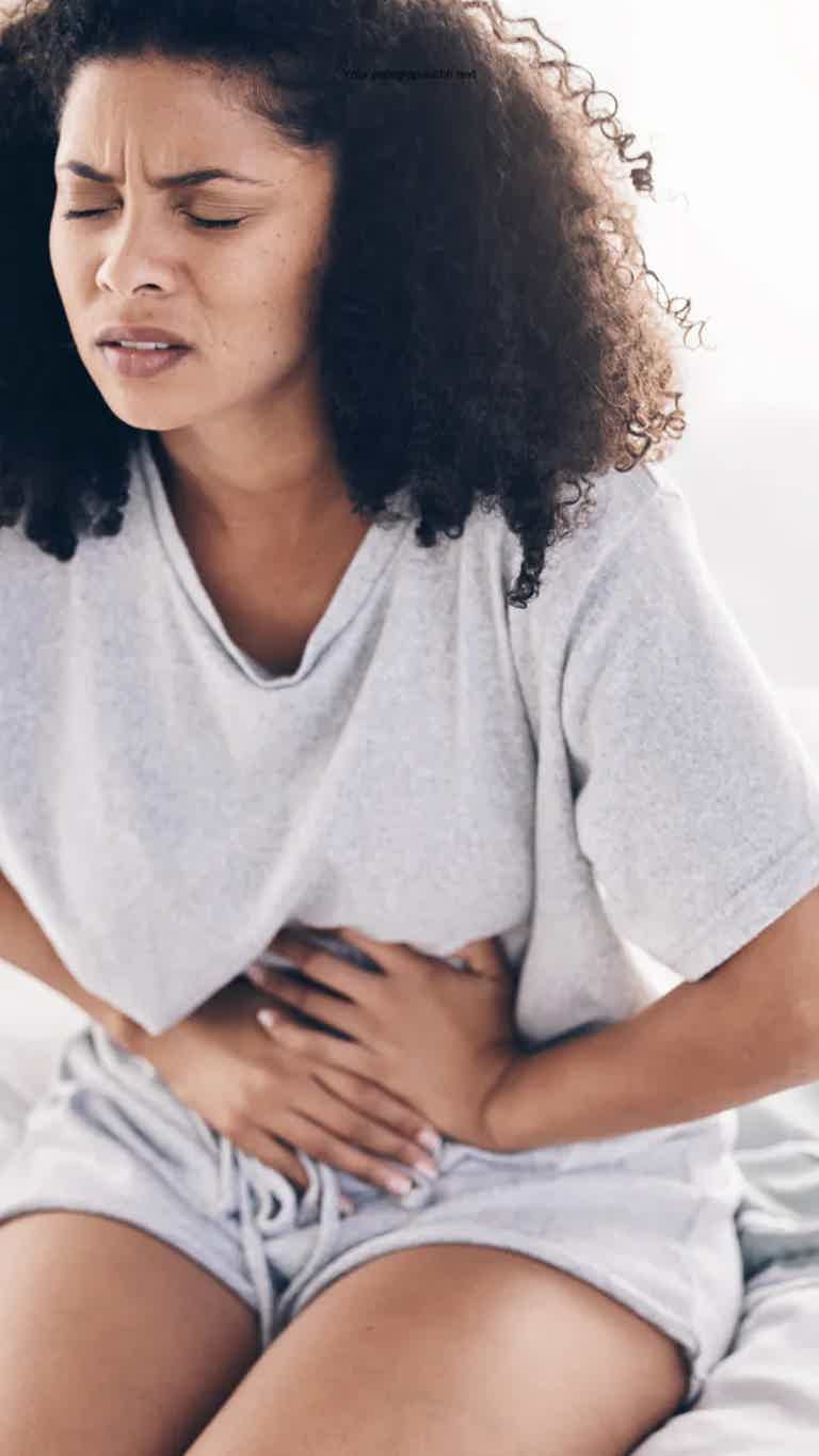 Woman clutching stomach in discomfort, eyes closed, seated in light gray clothing.