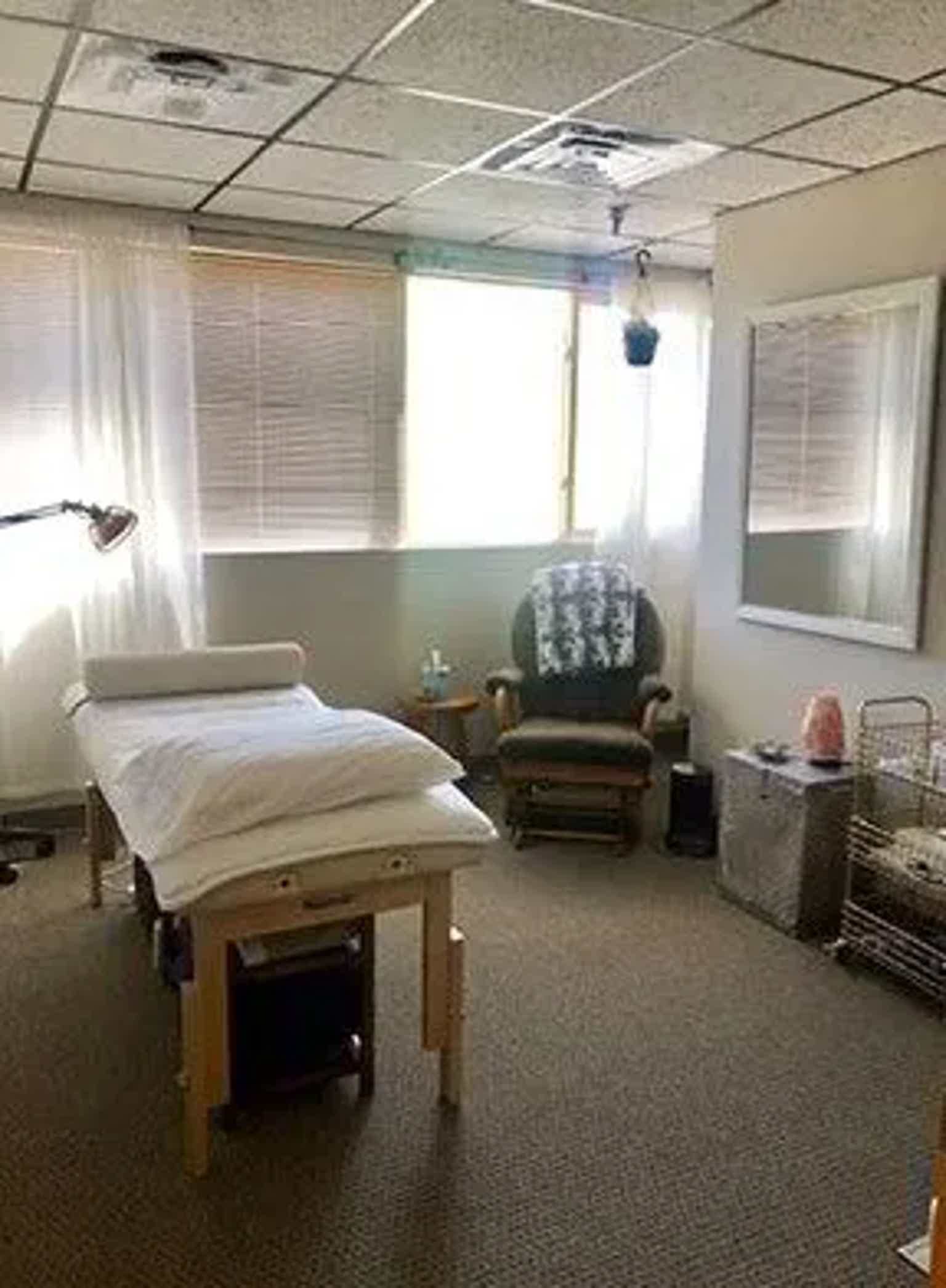 Clinical exam room with treatment table, chair, cabinets, window blinds, and overhead lighting.