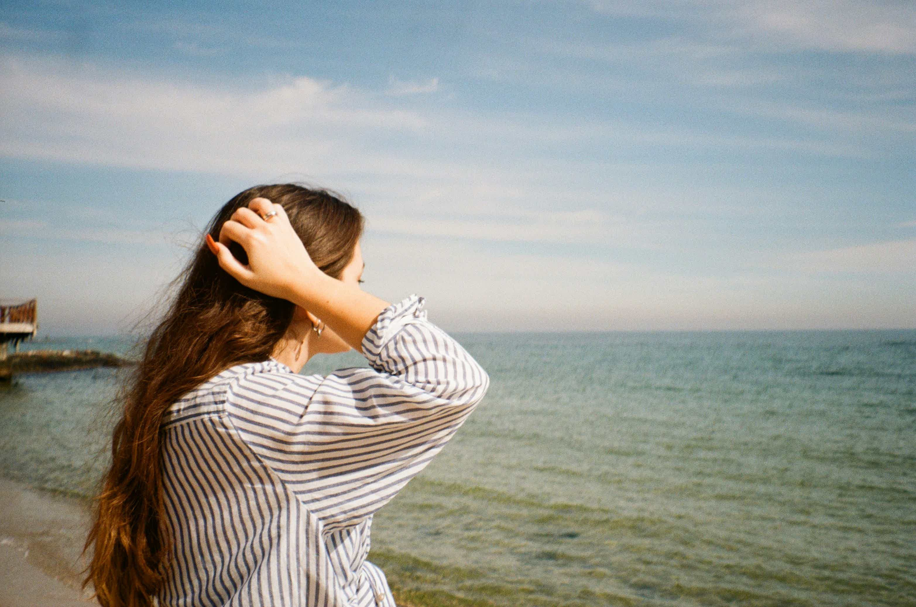 Woman with long hair in striped shirt covering her eyes at the beach, looking toward the ocean under a blue sky.