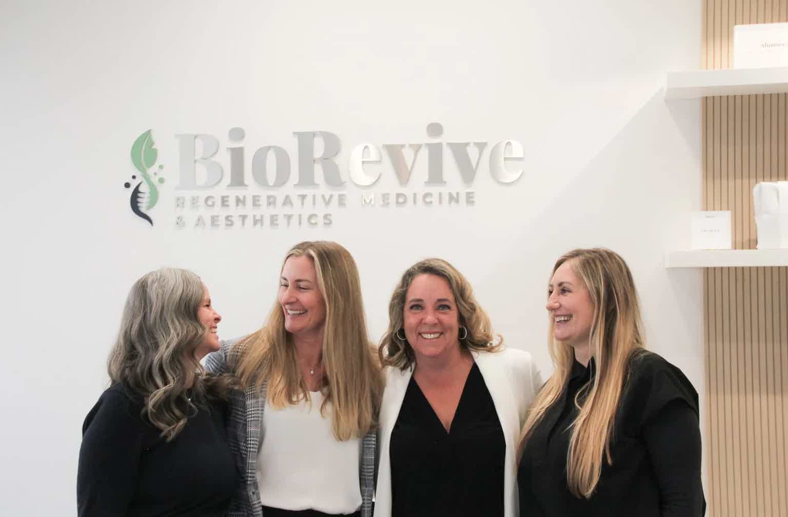 Group of four women smiling and chatting in a clinic lobby with BioRevive logo in background.
