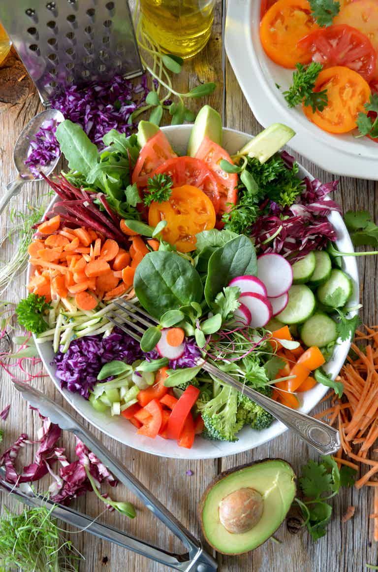 Colorful fresh salad with lettuce, cucumber, tomatoes, carrots, radish, avocado, kale, and herbs on a wooden table.