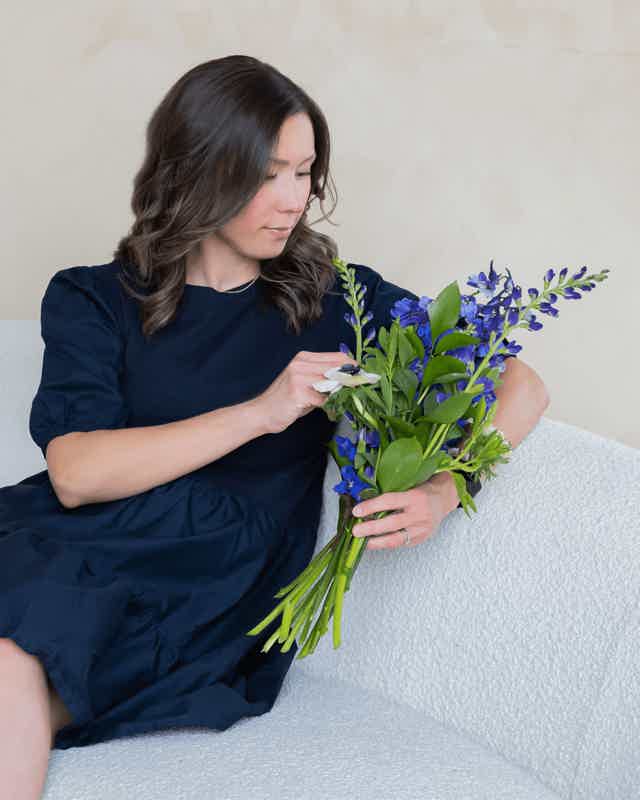 Woman in navy dress sits on sofa, smelling a bouquet of bluebells and greenery.