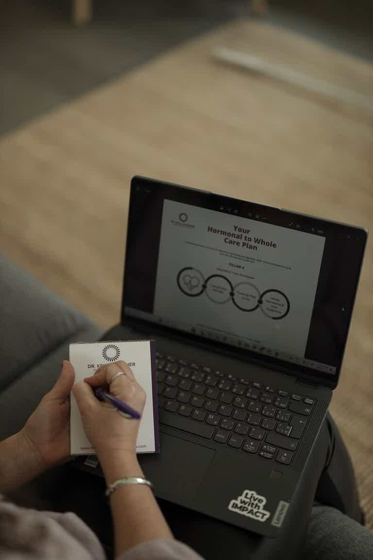 Person writing on notepad in front of open laptop showing a presentation on screen.