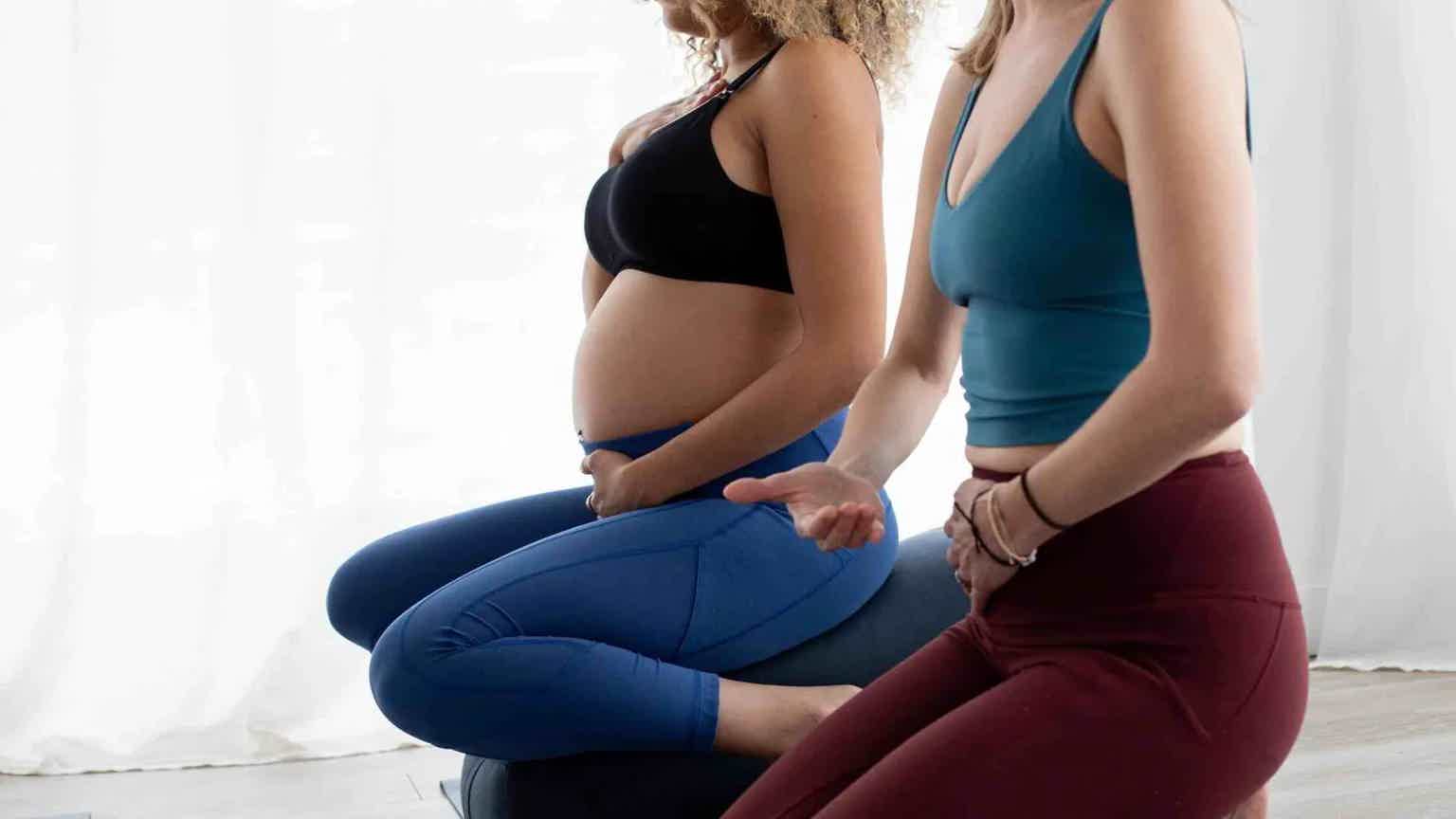 Two women in sports bras and leggings seated side-by-side, one with blue leggings, posed casually against a white backdrop.
