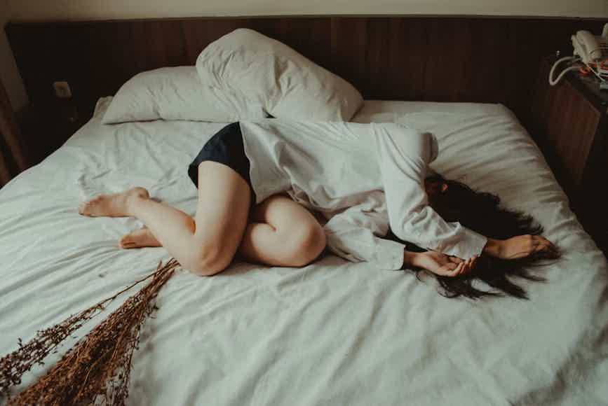 A woman lying on a white bed curled up to relieve her menstrual cycle pain.