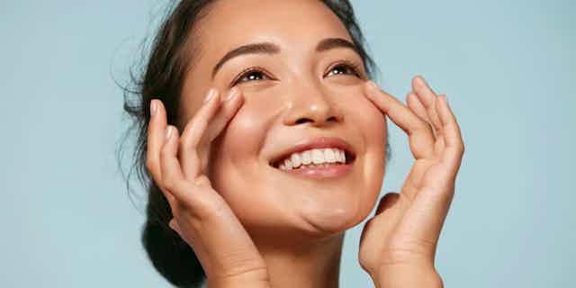 Smiling woman touching cheeks with hands, looking up, against light blue background.