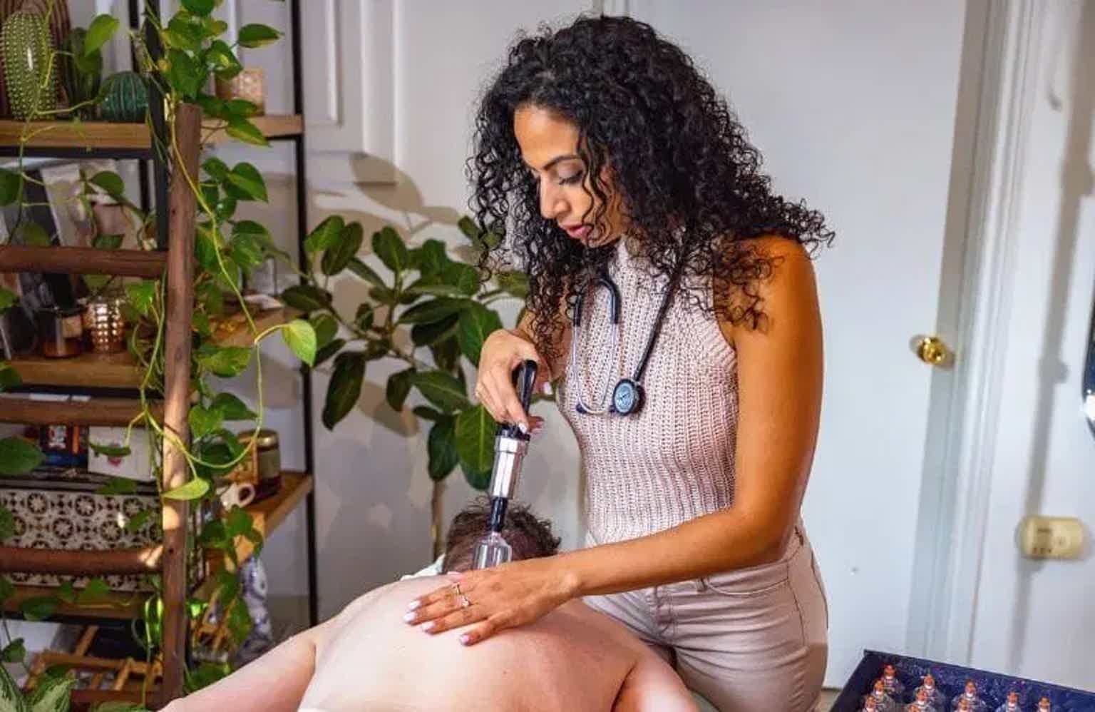 Woman with curly hair seated indoors, using a handheld device; plants and door in background.