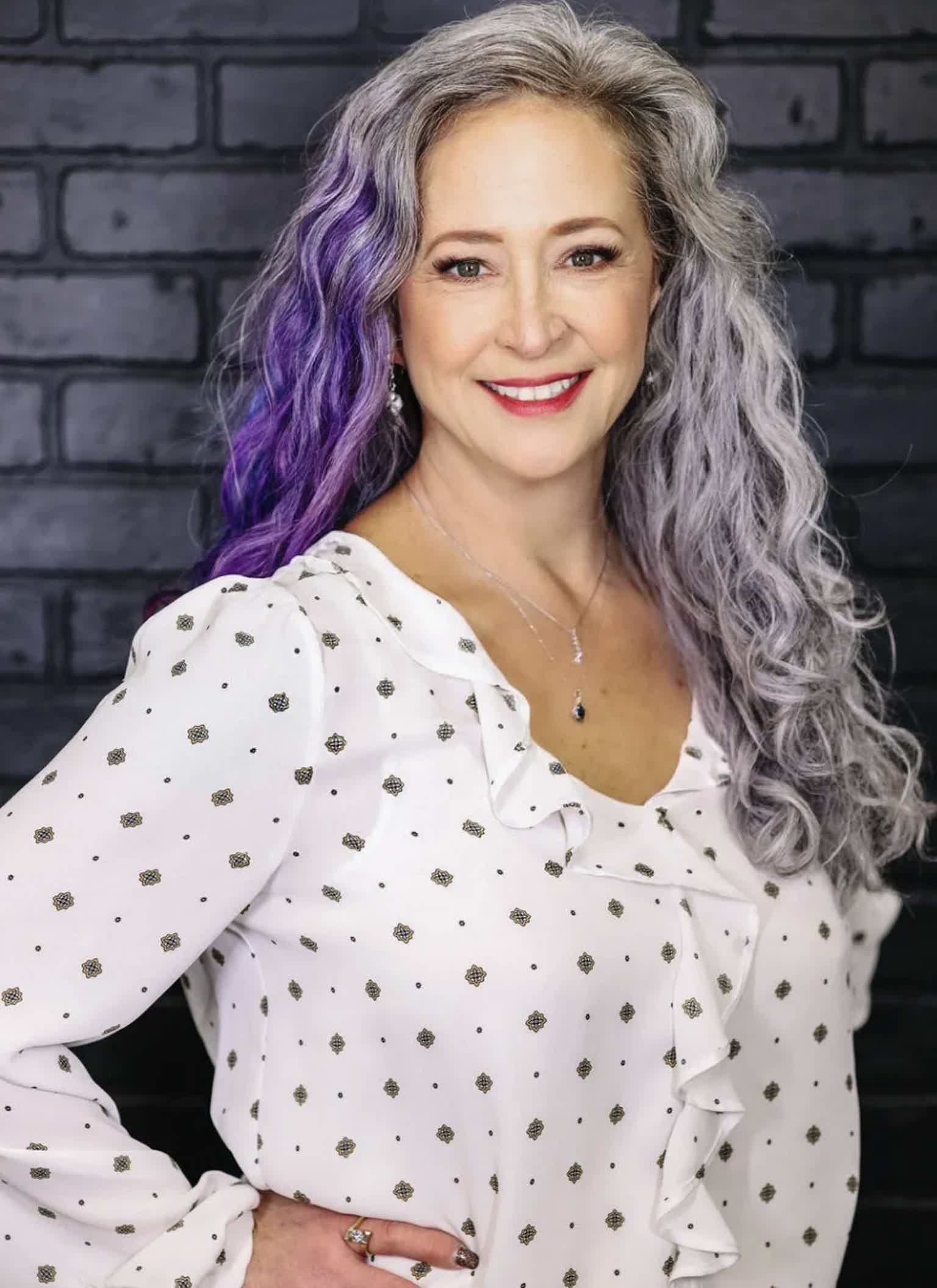 Smiling woman with wavy gray and purple hair, wearing a white polka-dot blouse with ruffles, against a dark brick wall.
