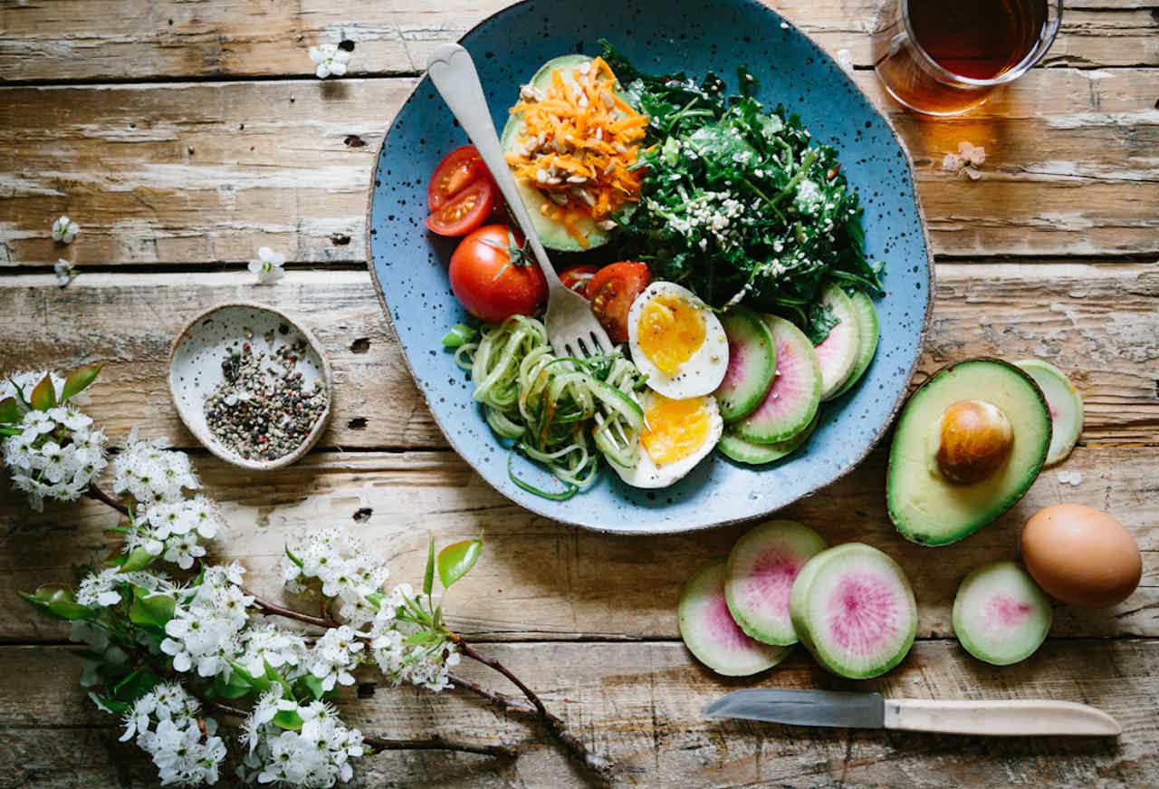 Colorful salad bowl with tomato, avocado, cucumber slices, poached eggs, and shredded carrots on rustic wooden table.