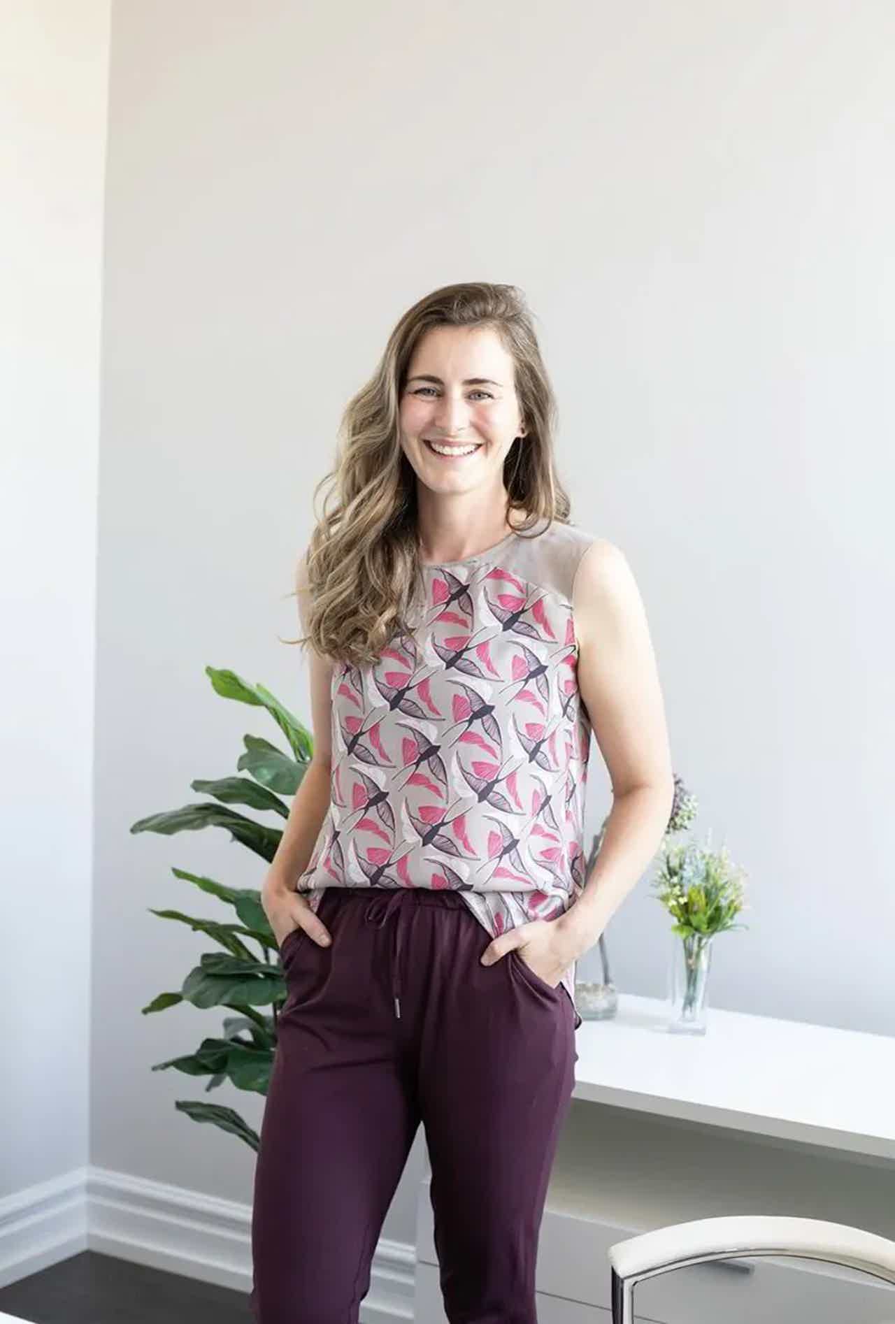 Smiling woman with curly hair in patterned sleeveless top and maroon pants, posing beside potted plants indoors.