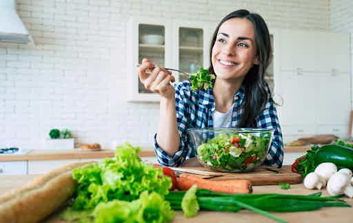 Woman happily tasting a fresh green salad at a kitchen counter surrounded by whole vegetables, illustrating a gut-healthy diet.