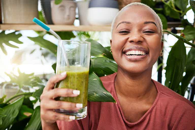 Smiling woman holds up a clear glass of green smoothie, surrounded by leafy plants in a bright indoor setting.