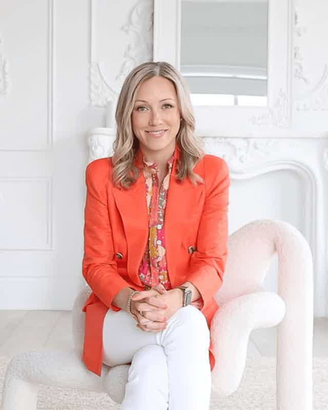 Woman sits on a fluffy white chair in a bright room, wearing a coral blazer and patterned blouse, smiling at camera.