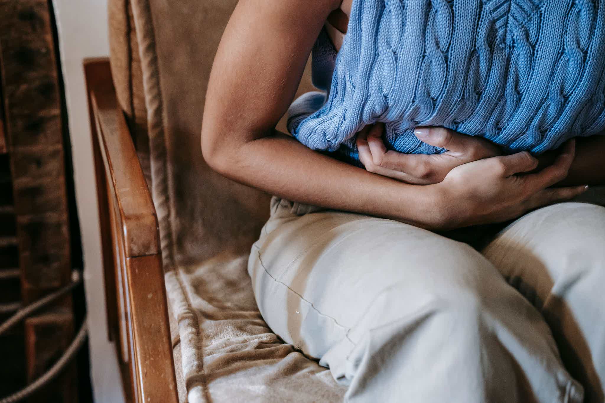 Person sits in a chair, arms wrapped around their midsection in apparent abdominal discomfort.