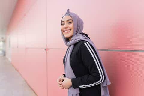 Smiling woman in black athletic jacket with gray hijab standing outdoors against pink metal wall.