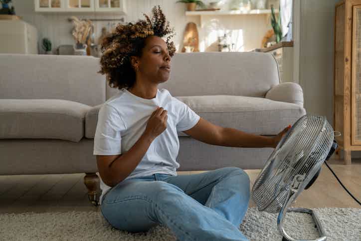 Woman in white T-shirt and jeans seated on living room carpet, holding a small drum-like object, smiling.