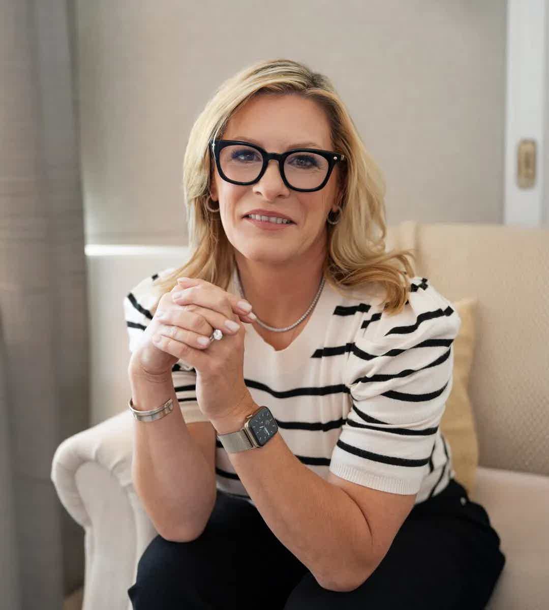 Smiling blonde woman wearing black glasses and a striped top, seated indoors and clasping hands on her knee.