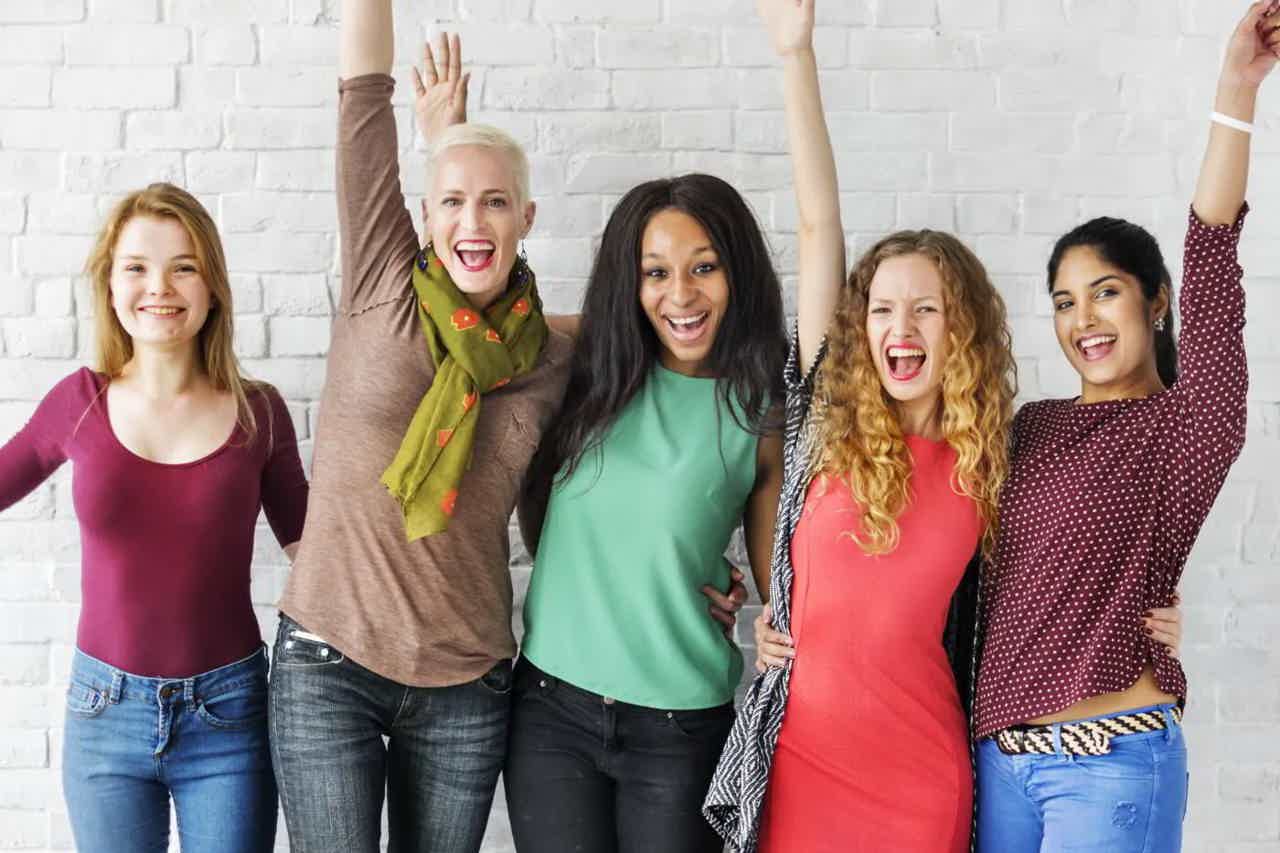 Group of five friends cheering with raised hands and bright smiles against a white brick wall.
