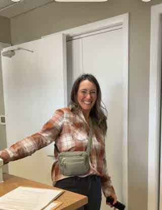 Smiling woman in a plaid shirt holding a pen at an office desk, with a white sliding partition behind her.