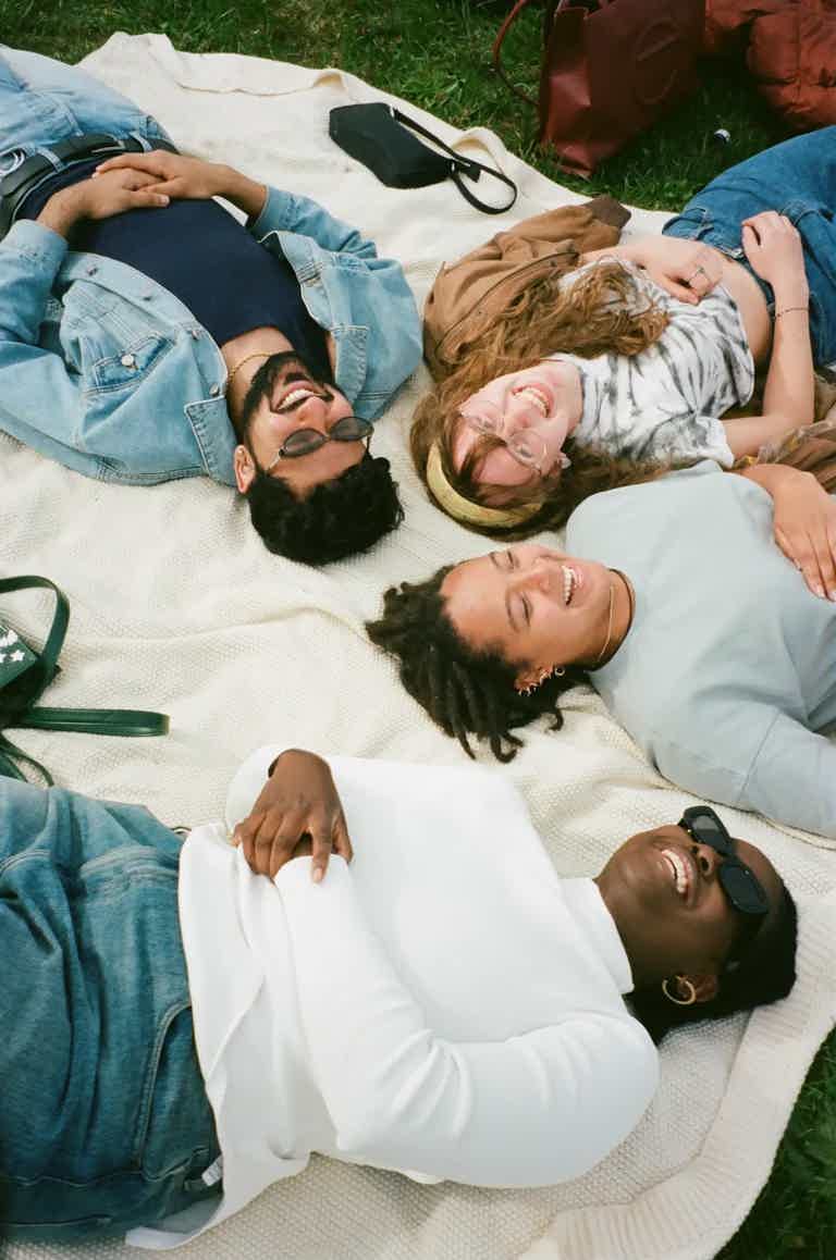 Group of five friends lying on a blanket in a park, smiling and relaxing together.