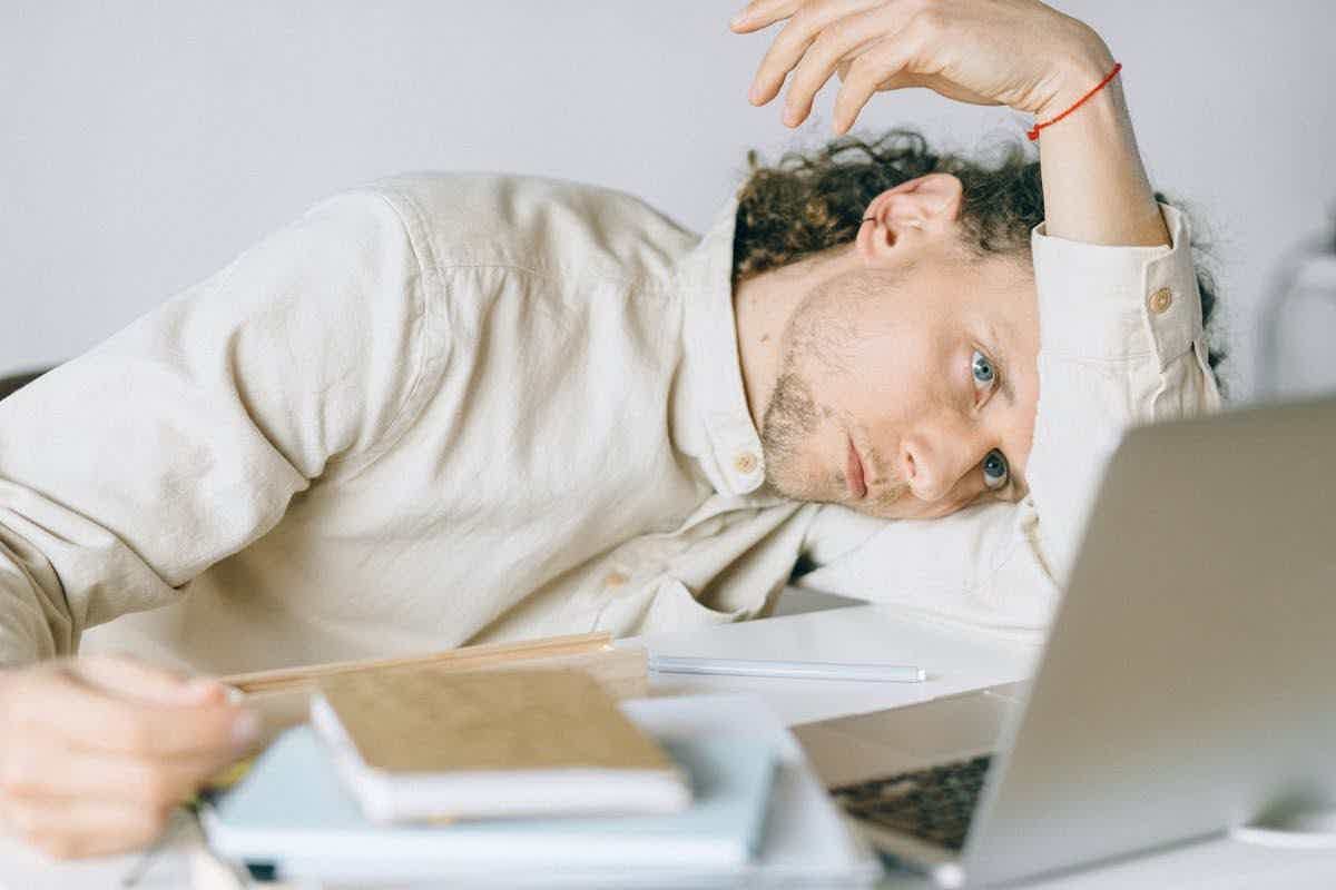 A man resting his head on his arm at a desk beside a laptop, looking drained and unable to focus.
