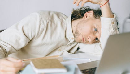 A man resting his head on his arm at a desk beside a laptop, looking drained and unable to focus.
