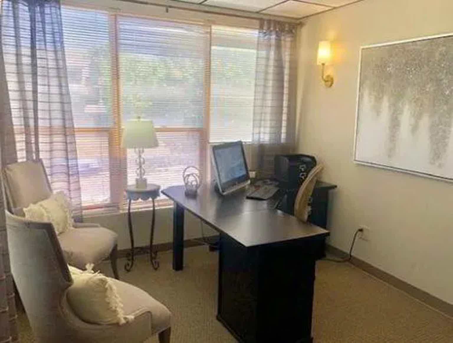 Bright office corner with two desks, computer monitors, chairs, and large window blinds with sheer curtains.