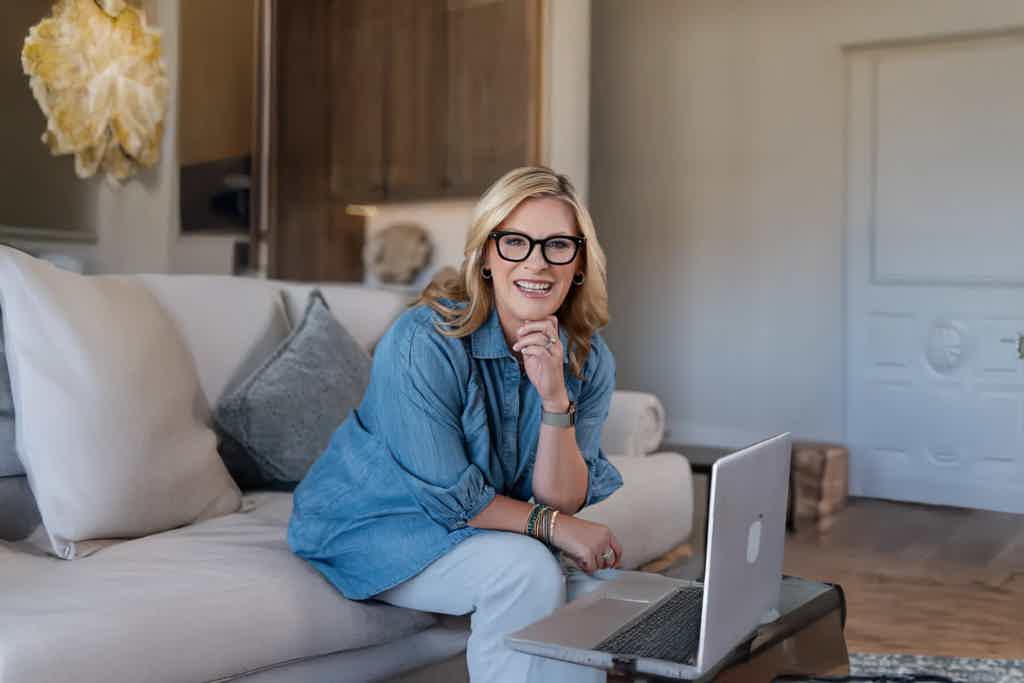 Smiling woman with glasses sits on a couch with laptop at a cozy home office.