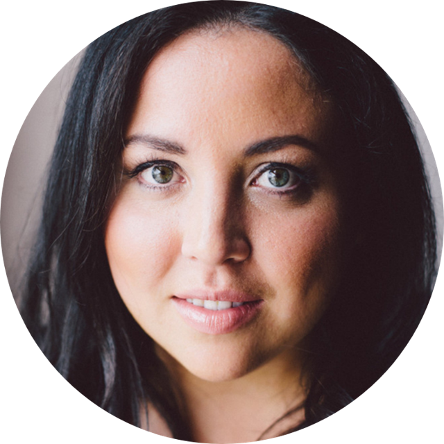 Smiling woman with blonde hair wearing a black wide-brim hat and black top, sitting against a dark backdrop.