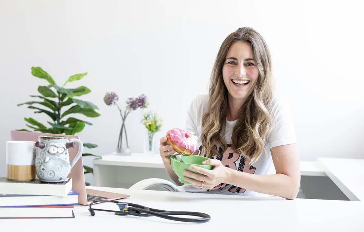 Smiling woman at desk holding a pink frosted donut over a green bowl, with laptop, books, plants, and stethoscope nearby.