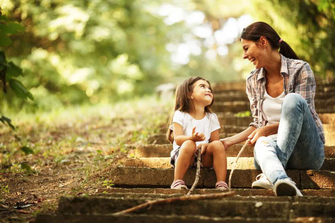 Mother and child sit on outdoor stone steps, smiling and talking in a sunlit garden.