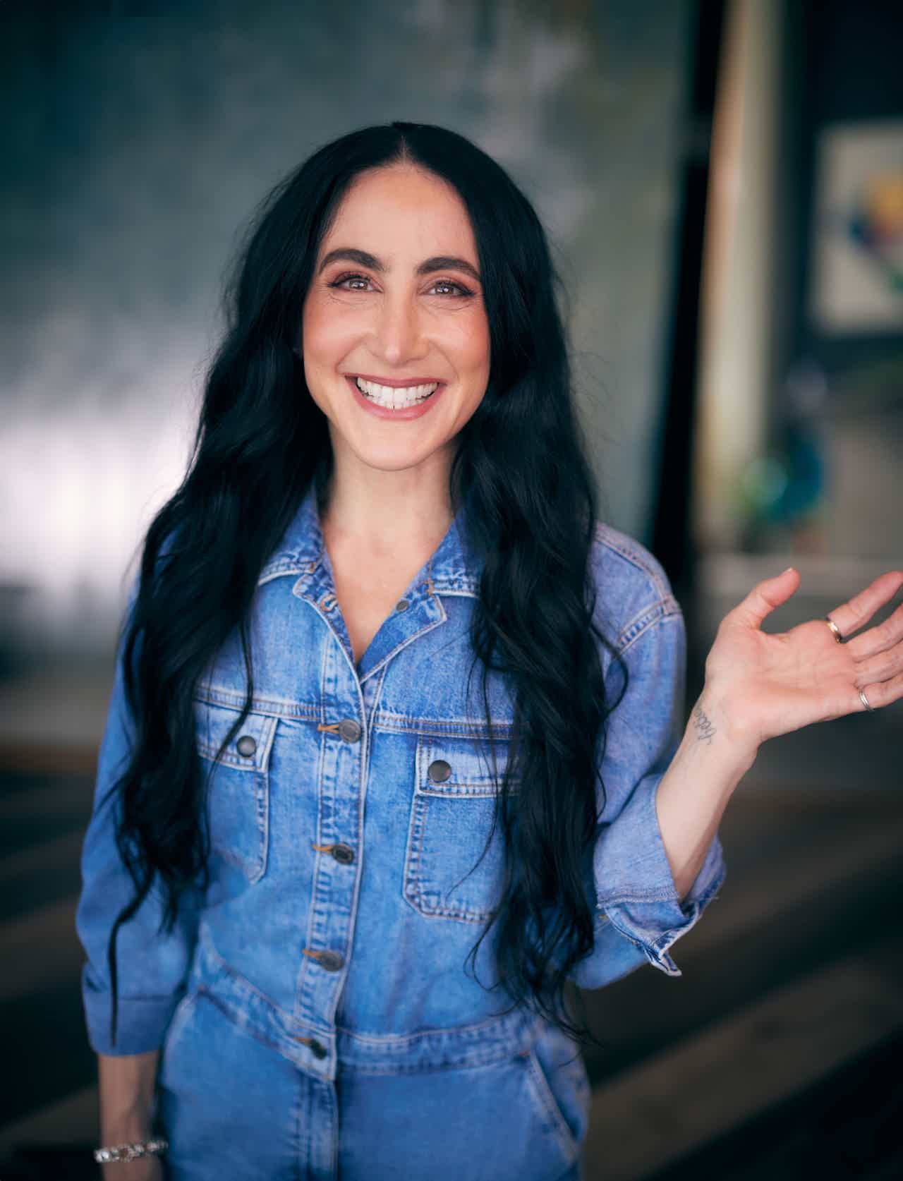 Smiling woman with long dark hair in a denim jacket waves her hand inside a studio.