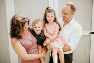 Family of four: mother and father holding two smiling children in front of light-colored fence.