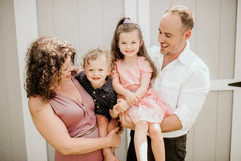 Family of four: mother and father holding two smiling children in front of light-colored fence.