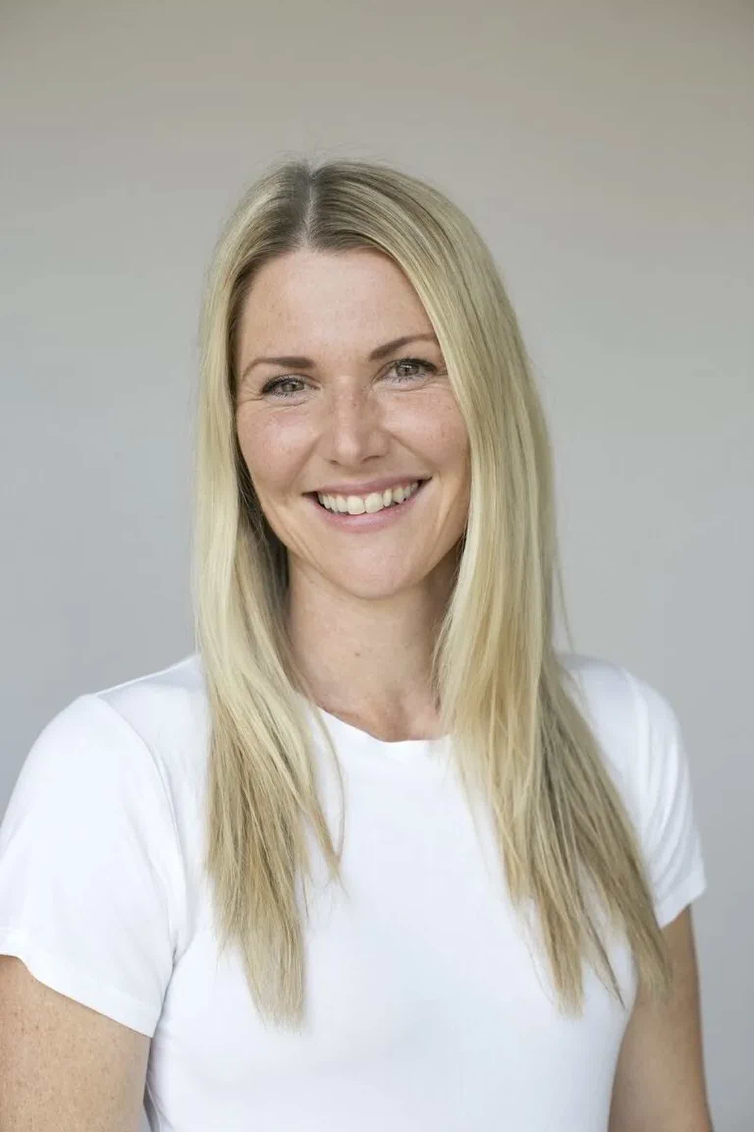 Smiling blonde woman with shoulder-length hair wearing a white top against a light gray background.