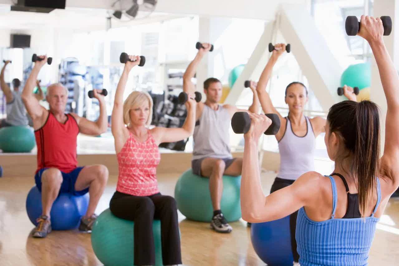 Group fitness class doing overhead dumbbell presses on stability balls in a bright gym.