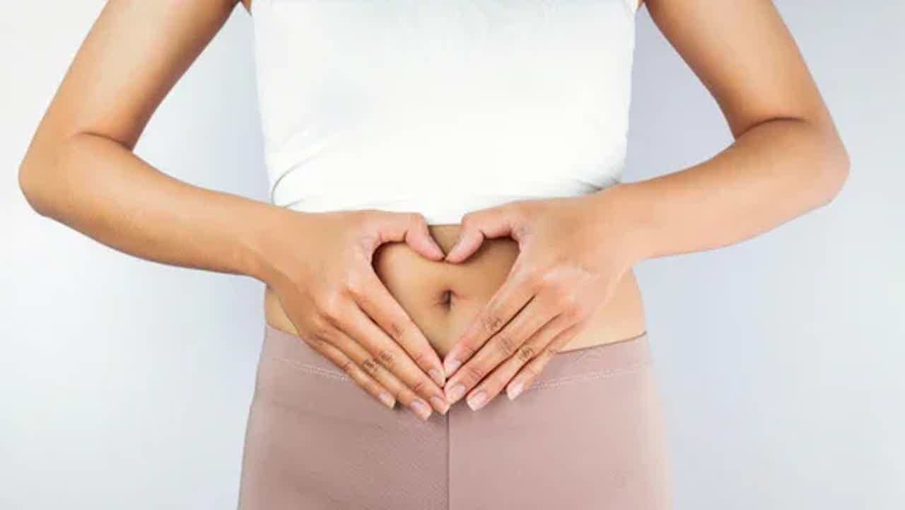 Person forming a heart shape with hands over bare midsection, wearing a white top and beige leggings.