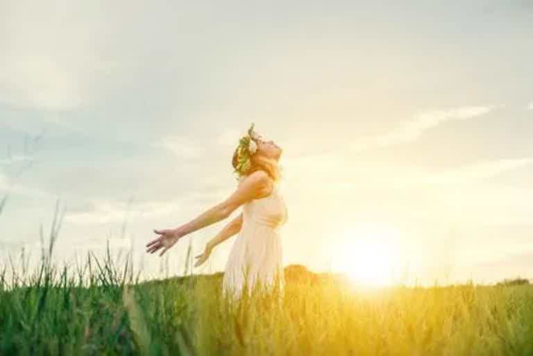 Woman in a white dress with flower crown stands in a sunlit field, arms outstretched, warm glow behind.