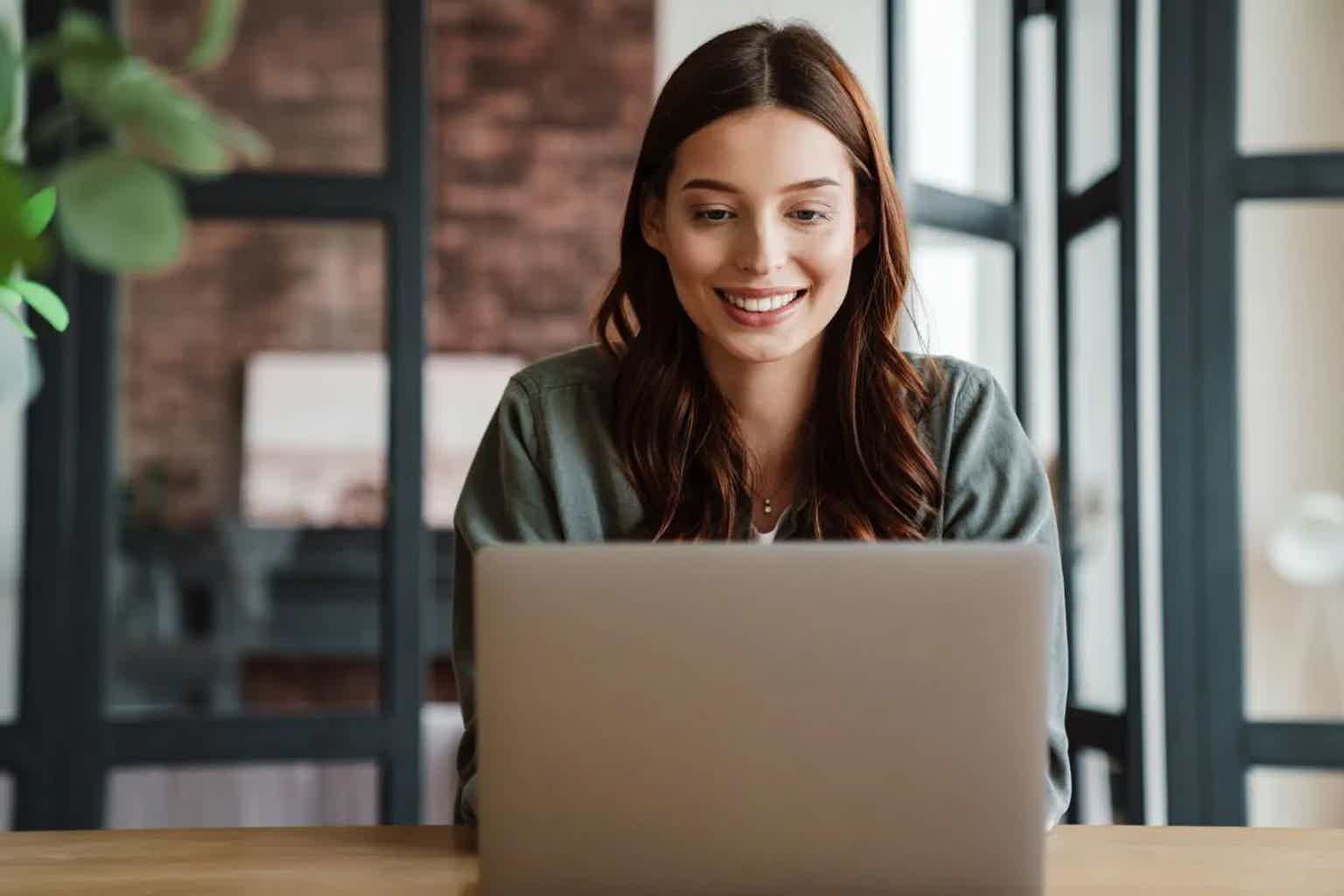 Woman smiling at a desk, working on a laptop in a bright office with window shutters behind her.