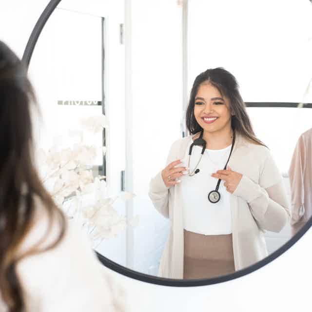 Smiling woman with long dark hair, white sweater, seated on pink chair, plant beside her in a bright room.