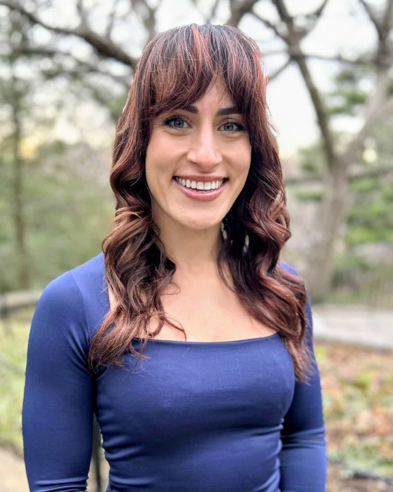 Smiling woman with long wavy red hair wearing a green top, outdoors with greenery in the background.